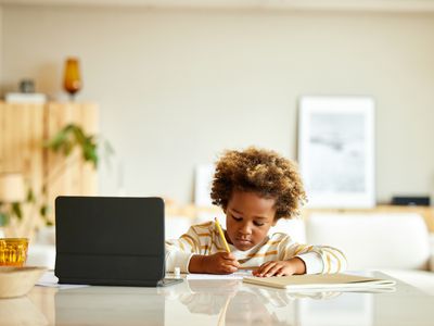 A child writing on paper with a tablet computer on the counter nearby.