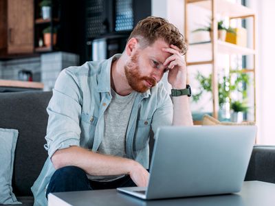 A man with red hair and a blue shirt looking annoyed at his laptop while sitting on a couch.