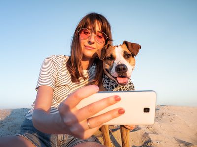 Low Angle View Of Young Woman Taking Selfie With Dog Through Smart Phone At Beach Against Clear Sky