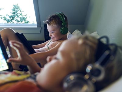 Two children laying on a bed with gaming devices in their hands and headphones on